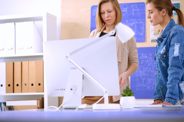 Two young woman standing near desk with instruments, plan and laptop.