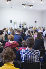 Obraz premium Group of People Sitting in hall During a Conference. Two Male Hosts Speaking on Stage in Front of the Audience.