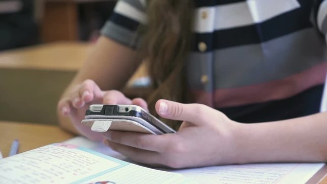 Schoolgirl dressed in a jacket sits at a school desk and holding a silver smartphone