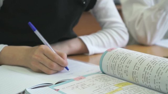 The schoolgirl sits at a school desk and writes in a notebook with a ballpoint pen. Close-up