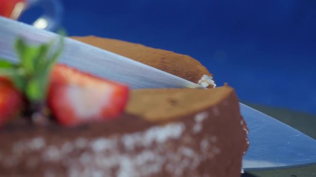 Female cutting chocolate cake with strawberries on wooden table, on dark blue background. Slice of glazed chocolate cake on spatula. Close up