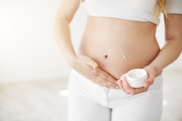 Close up of blonde early pregnant woman taking care of her belly skin using vitamin creme. Mother care concept. High key.