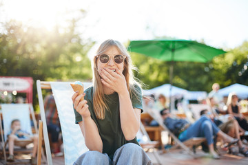 Lady eating ice cream laughing. Portrait of young female sitting in a park on a sunny day eating icecream looking at camera wearing glasses enjoying summer.