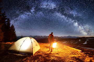 Male tourist have a rest in his camp near the forest at night. Man standing near campfire and tent under beautiful night sky full of stars and milky way, and enjoying night scene. Astrophotography - Powered by Adobe