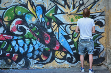 Photo of a young guy in denim shorts and a white shirt. The guy draws on the graffiti wall a...