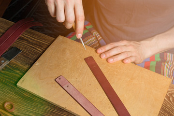 The master holds in his hand an awl and working with leather. On brown wooden table scattered with tools and accessories.