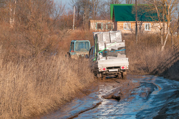 Wheeled tractor tows off-road truck with furniture settled down in mud on dirt road in village
