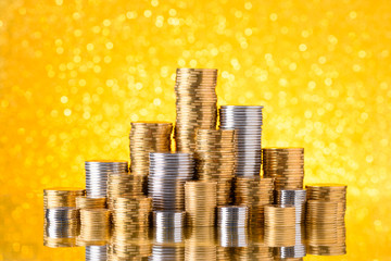 Shiny golden and silver coins stacked on gold glitter bokeh background with reflection
