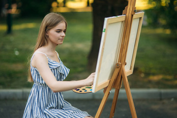 The blonde girl paints a painting on the canvas with the help of paints. A wooden easel keeps the picture.