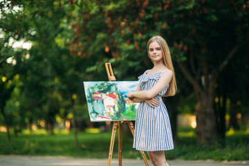 The blonde girl paints a painting on the canvas with the help of paints. A wooden easel keeps the...