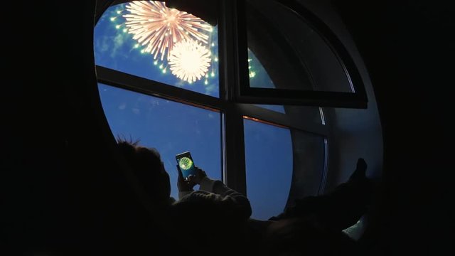 Girl taking fireworks video with a smartphone. Silhouette in a round loft window