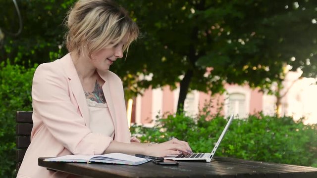 Attractive blonde white woman working freelance with laptop and smartphone in park at sunny day