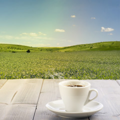 steamed cup of coffee on wooden table with sunny landscape on background.