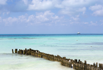 Seascape view on the tropical beach. Wooden breakwater on the shore. White yacht approaching the docks.