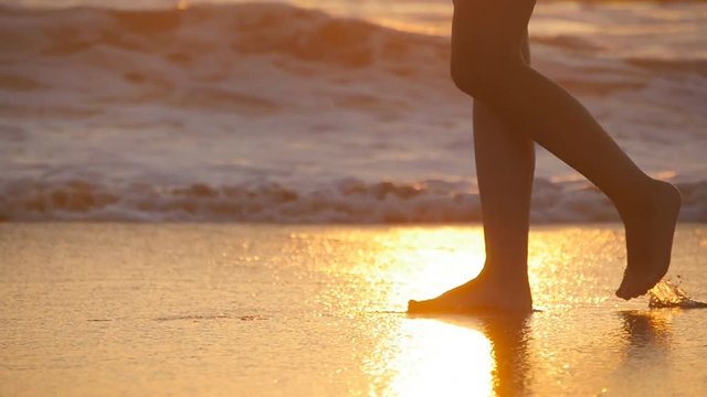 Female feet walking barefoot on sea shore at sunset. Legs of young woman going along ocean beach during sunrise. Girl stepping in shallow water at shoreline. Summer vacation. Slow motion Close up