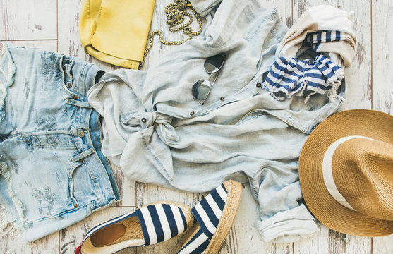 Summer outfit flatlay. Pastel summer women's clothes, parquet background, top view. Blue denim shorts, linen shirt, straw hat, yellow bag, sunglasses, blue and white striped neckerchief, espadrillas