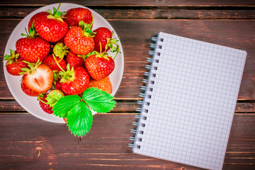fresh strawberries in bowl on  wooden background with notebook. Top view and copy space.