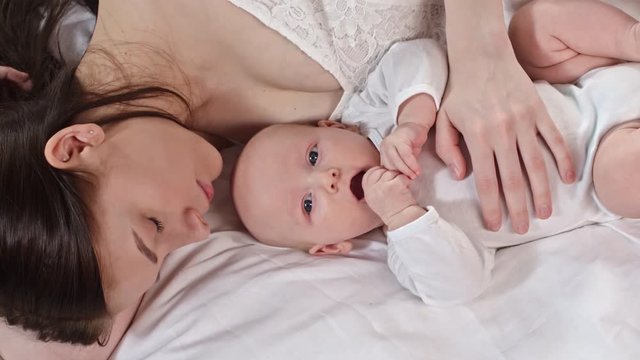 Top view of mother sleeping on bed and embracing little baby playing and putting his hand into mouth