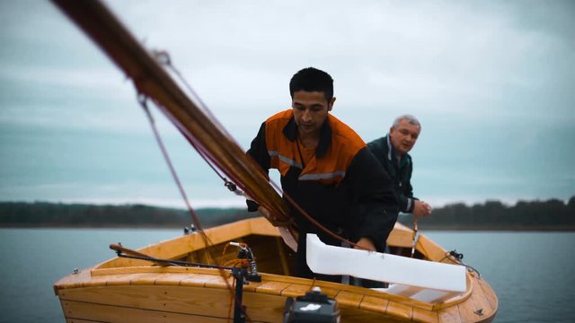 Wooden Sailboat in river with mast. Mature man author of boat and his assistant try to stand mast.
