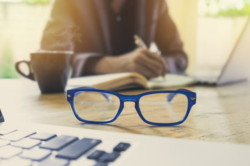 close up of spectacles and business man working and writing on notenook with laptop computer in blurred backfround. selective focus.