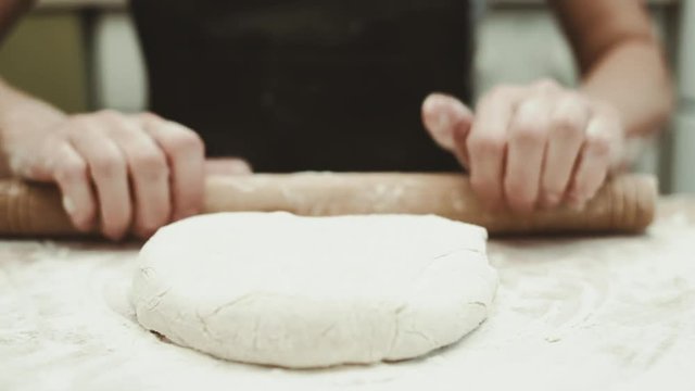 Woman in apron rolling raw dough on board. 