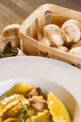 Fototapeta na wymiar Basket of raw mushrooms and white plate with freshly prepared pasta ravioli and fried champignons cut into slices. The dish is decorated with a sprig of rosemary. Traditional Italian homemade food.