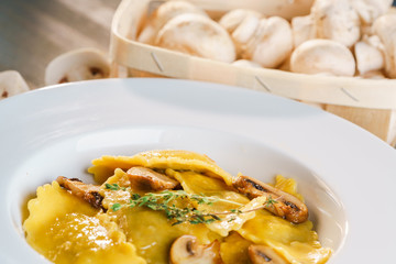 Basket of raw mushrooms and white plate with freshly prepared pasta ravioli and fried champignons cut into slices. The dish is decorated with a sprig of rosemary. Traditional Italian homemade food.