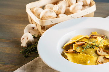 Basket of raw mushrooms and white plate with freshly prepared pasta ravioli and fried champignons cut into slices. The dish is decorated with a sprig of rosemary. Traditional Italian homemade food.