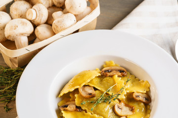 Basket of raw mushrooms and white plate with freshly prepared pasta ravioli and fried champignons cut into slices. The dish is decorated with a sprig of rosemary. Traditional Italian homemade food.