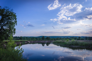 Calm beautiful scenic landscape with blue river, green trees and reflecting in water with cloudy sky. Magical sunset over the river in rural terrain. Natural, wild landscape.