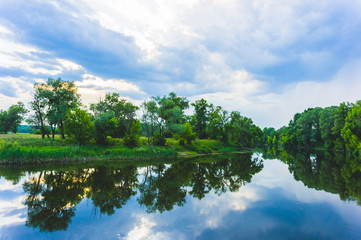 Fototapeta na wymiar Calm beautiful scenic landscape with blue river, green trees and reflecting in water with cloudy sky. Magical sunset over the river in rural terrain. Natural, wild landscape.