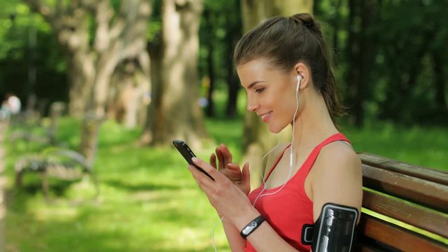 Hansome woman sitting on a bench and using smart phone, typing, texting, surfing internet, listening music at the park. Close up