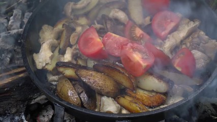 Frying potato with mushroom, tomato and onion in butter using a frying pan. Preparing food on campfire in wild camping.