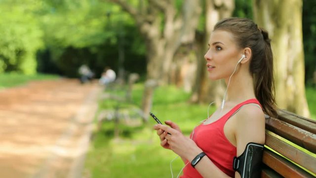 Attractive woman using smartphone while sitting on the bench. Fit woman looking at the runner.