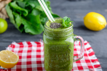 Healthy green spinach smoothie in a jar mug decorated with mint and chia seeds with ingredients on the checkered red napkin on the black wooden table. Selective focus