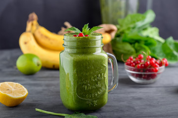 Healthy green spinach smoothie in a jar mug decorated with mint and red currant berries with ingredients on the black wooden table. Selective focus. Detox concept.