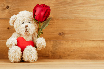Brown teddy bear with red heart and rose on wooden table.
