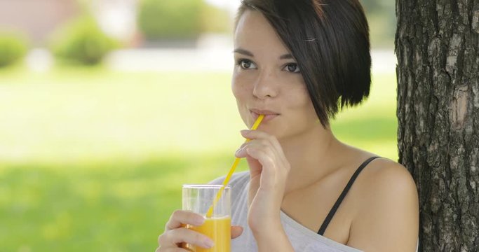 Young woman drinking fresh orange juice outdoors at summer park tree. Healthy lifestyle food.
