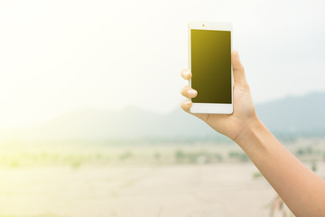 Female right hand holding and touching on mobile smartphone show back side. He is searching something. on blur forest mountain and sky background
