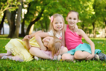 Naklejka na ściany i meble Three cute little sisters having fun together on the grass on a sunny summer day. Funny kids hanging together outdoors.