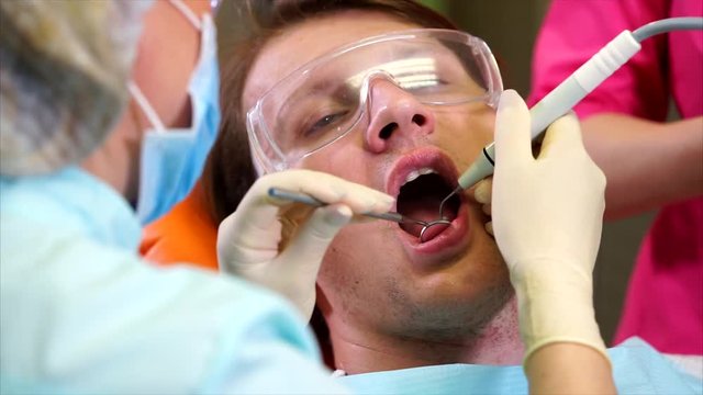 Dentist using mirror tool and scaler equipment to remove dental calculus from patient teeth, assistant helps with salivary ejector