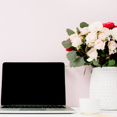 Home office desk with blank screen laptop, beautiful roses and eucalyptus bouquet in front of pale pastel pink background. Blog, website or social media concept .