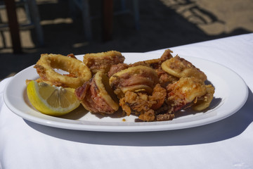 Fried squid rings served on a white dish by the beach.
Fried calamari portion with slice of lemon on white linen tablecloth in Greece.