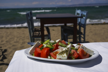 Greek salad with feta cheese, onions, olive oil and tomatoes by the sea.
Xoriatiki salad served on a table by Greek beach with blurred beach tavern table and wooden blue chairs background.