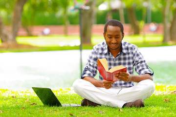 Young black tourist man sitting down on green grass reading a book with his computer near to him in the city of Quito Ecuador