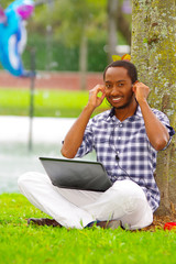 Young black man sitting down on green grass and working in his computer and listening music with his headphones posing his back in a tree in the city of Quito Ecuador
