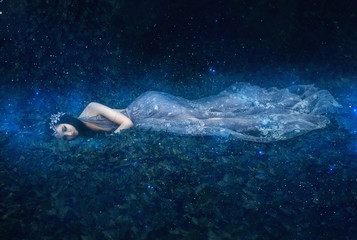 Fototapeta na wymiar fantasy woman Sleeping Beauty. girl lies on the grass in dark, dense forest magical woods. An unusual transparent blue creatuve long dress. Artistic processing art photography. Sweet dream