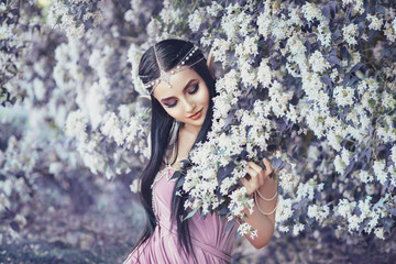 Portrait of an elf in a blooming garden. A girl with long ears touches the flowers. She is dressed...