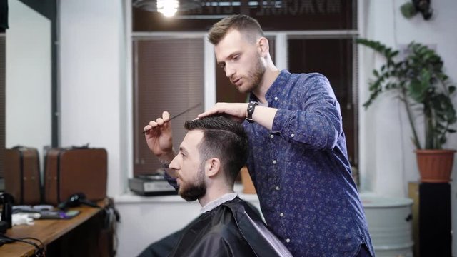 A stylish hairdresser gently combs his client's hair in order to create a stylish hairstyle in a beauty salon for the visitor with a beard