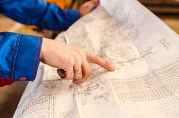 Naklejka premium The worker in overalls points with his index finger to the drawing of high-voltage poles. A man's hand against a background of a metallurgical plant close-up. Industrial Engineering.